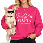 A woman wearing a bright pink sweatshirt with 'Tiny Dog MAFIA EST. 2025' printed in white, holding a small tan dog wearing black sunglasses.