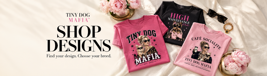 Three graphic T-shirts are arranged on a white fabric background with decorative flowers, sunglasses, and jewelry. The shirts feature humorous dog-themed designs: a bright pink 'TINY DOG MAFIA' shirt, a black 'HIGH MAJESTIC' shirt, and a light pink 'CAFE SOCIALITE' shirt.