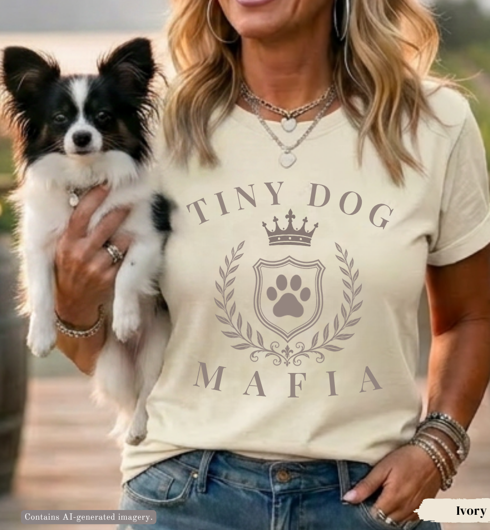 A woman wearing an ivory graphic t-shirt with a 'TINY DOG MAFIA' crown and paw print design, holding a small black and white Papillon dog in an outdoor setting.